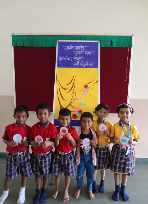 Children holding rosette greeting cards in front of Guru Purnima display